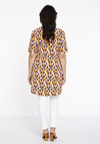 Tunique colorée avec un motif en losange orange, violet et rose, à manches courtes et à coupe droite, portée sur un pantalon blanc et des sandales à talons.