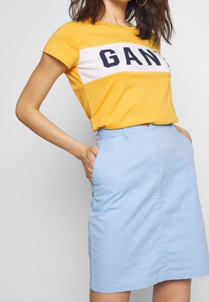 Woman wearing a yellow short-sleeve shirt with "GANT" text and a light blue knee-length skirt with hands in pockets.