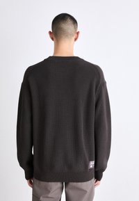 Maglione a maglia marrone scuro con texture a costine, scollatura a girocollo e spalle scese. Presenta una piccola toppa con logo sul lato inferiore della schiena.