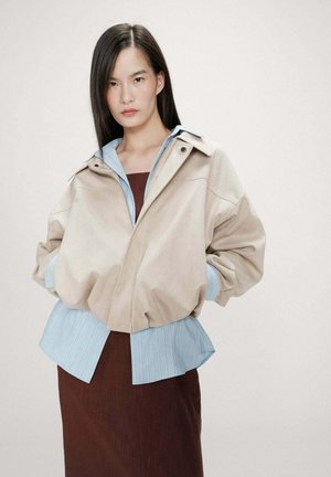 Jeune femme aux cheveux raides et foncés portant une veste beige sur une chemise bleue et une robe marron, debout devant un fond uni.