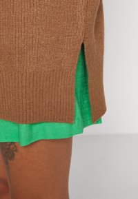 Brun strukturerad stickad tröja med sidospalt, lagd över en grön linneklänning, som visar upp kontrasterande färger och tygtyper.