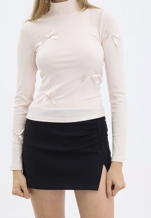 Hellrosa geripptes Langarmoberteil mit hohem Kragen und kleinen Satin-Schleifen. Kombiniert mit einem figurbetonten schwarzen Minirock mit seitlichen Raffung.