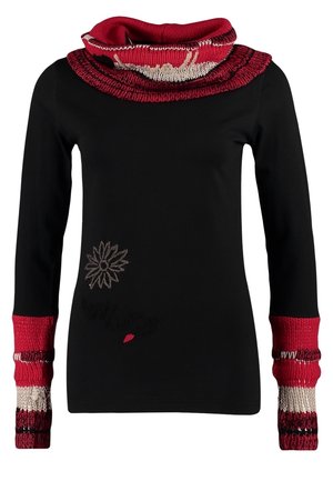 Vrouw draagt een zwarte top met rode en witte gebreide manchetten en kraag, blauwe spijkerbroek met ritsdetail, zwarte laarzen, houdt een zwarte clutch vast.