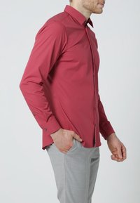 Rotes Langarmshirt mit schmaler Passform, mittlerem Kragen und dunklen Knöpfen, kombiniert mit hellgrauer Hose. Glatte Textur, keine Muster.