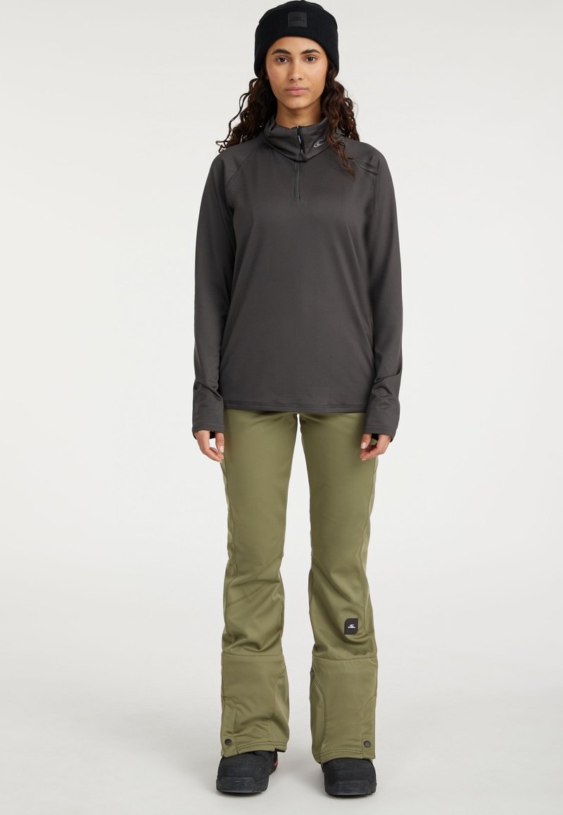 Langärmliges, dunkelgraues Oberteil mit halbem Reißverschluss, kombiniert mit olivgrünen Hosen mit weit geschnittenen Beinen und einem schwarzen Logo-Patch, getragen mit schwarzen Stiefeln.