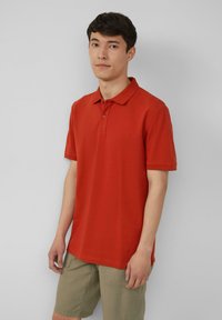 Rode polo shirt van katoen, met een klassieke kraag en korte mouwen. Model draagt het met licht kaki shorts. Subtiele logo detail op de borst.
