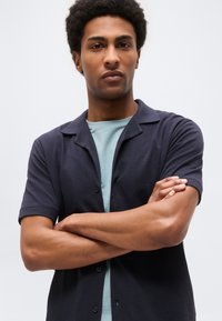 Ung mann med armene i kors, iført en mørk kortermet skjorte over en lys blå T-skjorte, ser selvsikkert fremover mot en ensfarget bakgrunn.