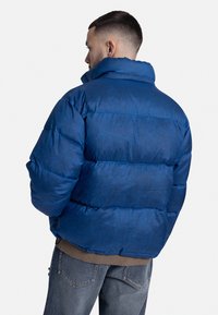 Modrá puffa bunda s vysokým golierom, prešívaným vzorom a hrubým polstrovaním. Textúrovaná tkanina a lamovaný spodný lem viditeľné zozadu.