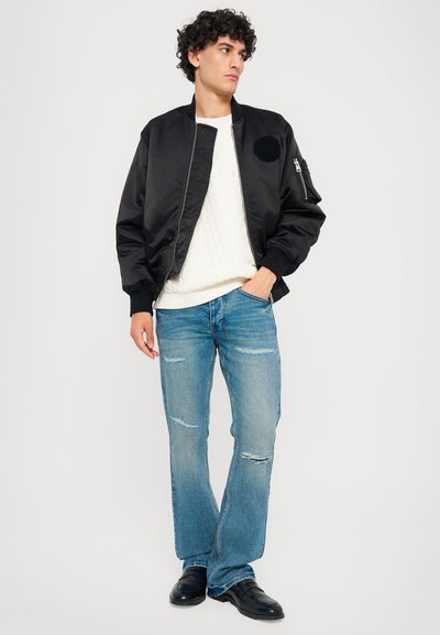 Μαύρο μπουφάν bomber με λείο φινίρισμα, ριμπ μανίκια και στρογγυλή στάμπα στον αγκώνα, συνδυασμένο με ανοιχτό μπλε τζιν αποσπασμένα και μαύρα παπούτσια.