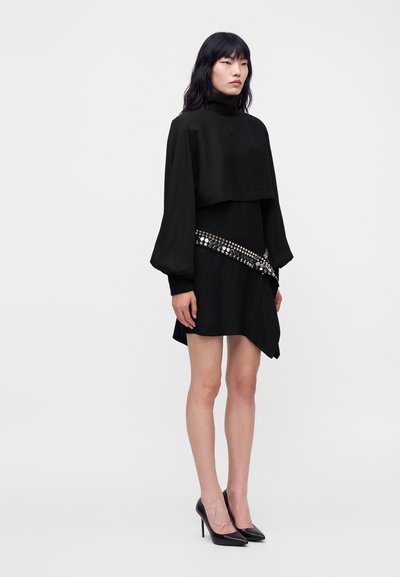 Mannequin portant une robe noire à col haut avec des manches volumineuses, une ceinture ornée et des talons noirs, debout devant un fond clair uni.
