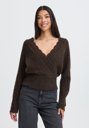 Femme souriante, portant un pull brun foncé à encolure en V ondulée et un jean foncé, debout devant un fond clair uni.
