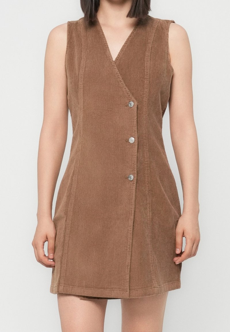 Robe en velours côtelé marron sans manches avec un col en V et trois boutons argentés sur le devant, portée par une personne debout devant un fond clair.