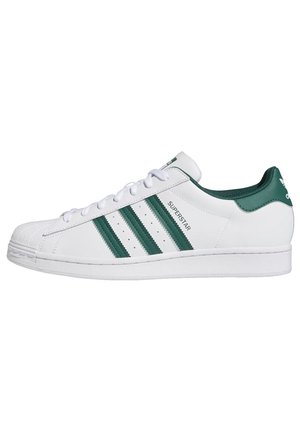 Biały sneaker Adidas Superstar z zielonymi trzema paskami, napisem „Superstar” i zielonym językiem na pięcie, pokazany z profilu.
