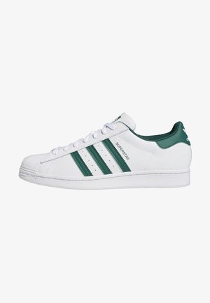 Biały sneaker Adidas Superstar z zielonymi trzema paskami, napisem „Superstar” i zielonym językiem na pięcie, pokazany z profilu.