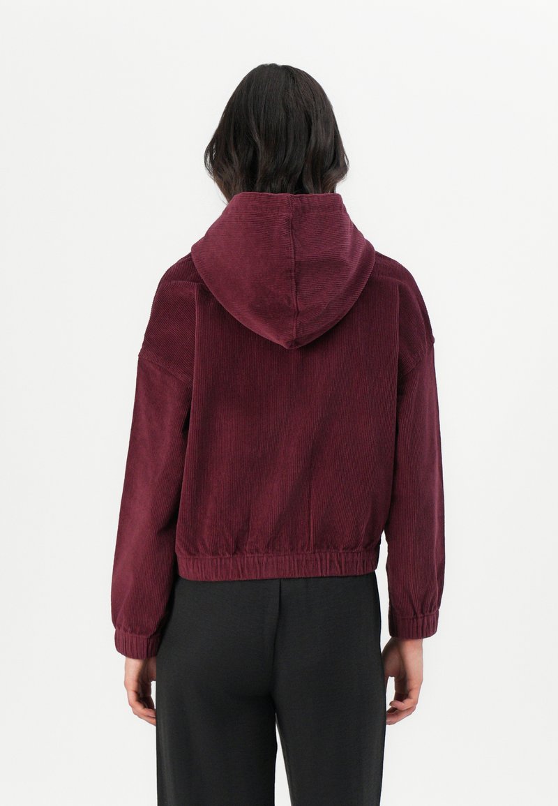 Bordowy, kordurowy, kapturowy płaszcz z zebranym dołem, o luźnym kroju i długich rękawach, pokazany od tyłu. Widoczna faktura.