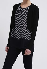 Cardigan nero sopra una maglia grigia con motivo a zigzag, abbinato a pantaloni scuri. Il tessuto sembra morbido con una vestibilità rilassata. Design semplice.