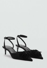 Zapatos de tacón alto de ante negro con punta afilada, que presentan un detalle decorativo de flecos en la parte delantera y una delgada correa en el tobillo.