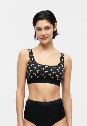 Woman with short dark hair wearing a black crop top with beige crescent moon patterns and high-waisted black bottoms against a plain background.