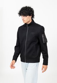 Zwarte zip-up jack met een hoge kraag en geribbelde manchetten. Heeft een logo op de borst en een zijzak met ritssluiting. Textuurstof.