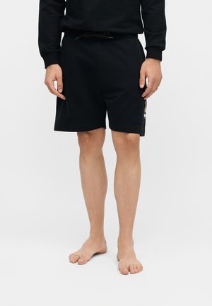 Άντρας που φοράει μαύρα σορτς έως το γόνατο και μαύρο μακρυμάνικο τοπ, όρθιος ξυπόλητος μπροστά σε απλό φωτεινό φόντο.