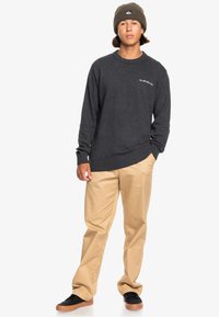 Grå tröja med ribbad nederkant och ärmslut, med en liten logotyp på bröstet. Beiga byxor med avslappnad passform och svarta sneakers fulländar looken.