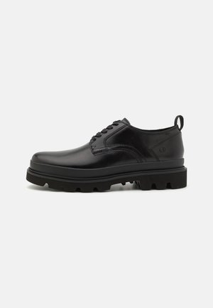 Chaussure noire en cuir à lacets avec semelle épaisse et robuste et languette au talon, vue de côté sur un fond clair uni.