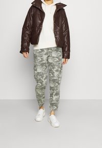 Brązowa kurtka puchowa z wysokim kołnierzem, jasnoszara bluza z kapturem pod spodem oraz zielone spodni cargo w kamuflażu, zestawione z białymi sneakersami.