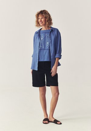 Femme portant une chemise bleue boutonnée par-dessus un t-shirt bleu imprimé de poissons, un short noir longueur genou et des sandales noires à enfiler, debout et souriante.