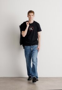 Czarna koszulka z logo, jasnoniebieskie dżinsy o prostym kroju i czarne buty. Model trzyma czarną kurtkę na ramieniu. Jednolity tle.