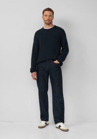 Mornarsko modra pletena pulover, temno modri džins in bele superge s rjavimi poudarki. Model stoji ob enobarvni sivi ozadju.