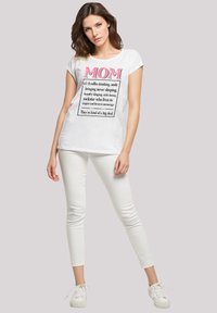 Weißes T-Shirt mit schwarzem Text und pinken Akzenten, auf dem "MOM" steht. Kombiniert mit weißen, eng anliegenden Hosen und weißen Sneakern. Einfaches, entspanntes Design.