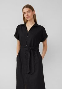 Abito nero con bottoni realizzato in lino leggero. Presenta un colletto, maniche arrotolate, cintura in vita, tasche e uno spacco per facilitare i movimenti.