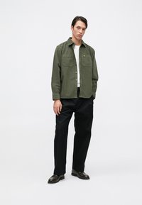 Camisa verde de manga larga con dos bolsillos en el pecho y un logo amarillo, combinada con pantalones negros y zapatos oscuros, sobre un fondo blanco.