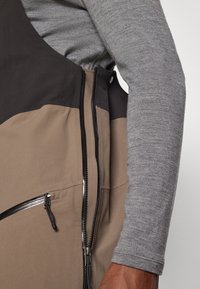 Uski blizu prikazuje prsluk s dva tona, smeđi i crni, sa patentnim zatvaračem, nošen preko sive košulje dugih rukava na ruci i torzu osobe.