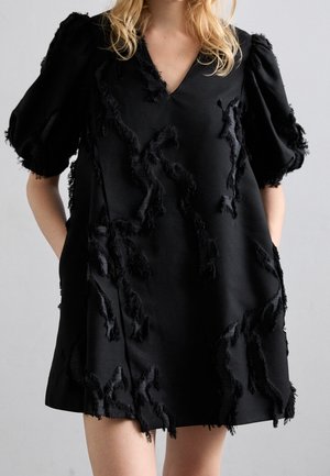 Frau trägt ein kurzes schwarzes Kleid mit Puffärmeln und strukturierten Fransen-Details auf dem Stoff, die Hände in den Taschen.