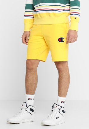 Man draagt felgele Champion shorts, witte FILA sokken, witte FILA sneakers en een meerkleurige gestreepte trui.