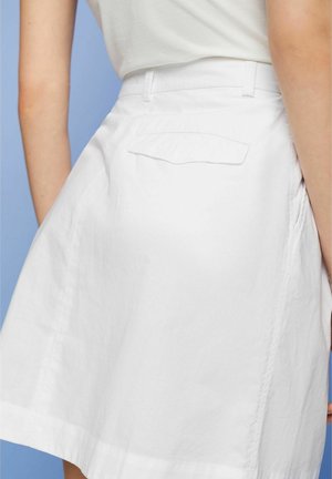 Jupe en coton blanc avec une poche à l'arrière, des détails de couture visibles et un ourlet droit, présentée sur un fond bleu.