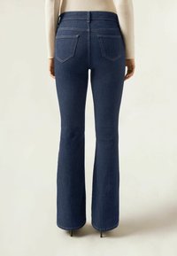 Mörkblå denimbyxor med flare och hög midja, två bakfickor och synlig sömnad; raka ben som smalnar av mot fållen.