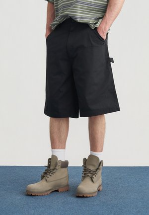 Pantaloncini neri in tessuto resistente, dotati di tasche laterali e taglio dritto. Abbinati a stivali beige con lacci e calzini bianchi a coste.