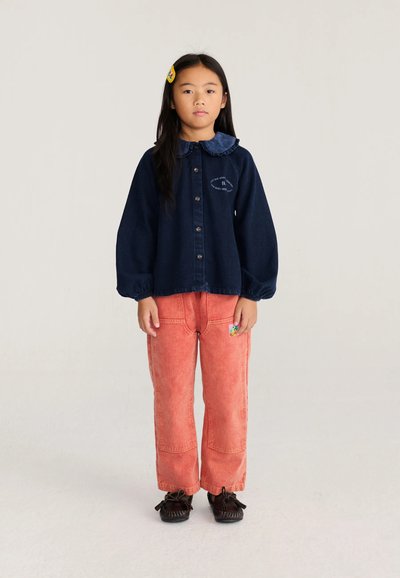 Jeune fille aux longs cheveux foncés portant une chemise bleu marine boutonnée, un pantalon corail, des chaussures foncées et une barrette colorée, se tient devant un fond uni.