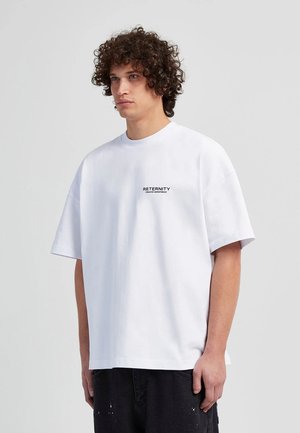 Weißes, übergroßes T-Shirt mit rundem Ausschnitt und kurzen Ärmeln. Verfügt über schwarzen Drucktext auf der Brust. Das Material wirkt glatt und leicht.