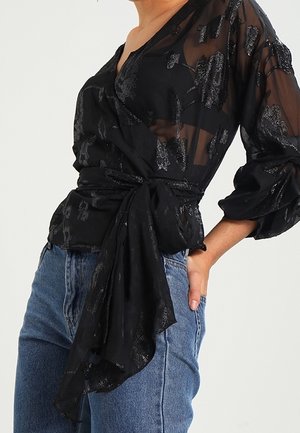 Personne porte un chemisier noir transparent à motifs métalliques, manches longues et ceinture à nouer, assorti à un jean bleu, main dans la poche.