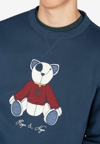 Sudadera de color navy con un diseño de oso bordado, que presenta un suéter rojo, acentos azules y detalles en blanco. El texto dice "Harper & Neyer."