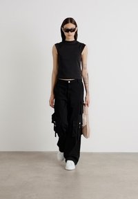 Black cropped sleeveless top, black cargo pants with pockets, white sneakers, and a light pink shoulder bag. Smooth texture, modern design.