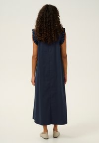 Mörkblå ärmlös maxiklänning med volangdetalj på axlarna och rak silhuett, tillverkad av ett mjukt tyg. Bärs med ljusa skor.