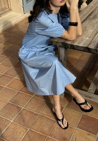 Femme portant une robe bleu clair et des sandales noires assise à une table en bois rustique sur un sol carrelé en terre cuite, les bras reposant sur la table.