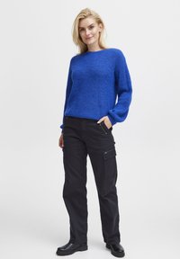 Maglione blu lavorato a maglia con maniche lunghe, abbinato a pantaloni cargo neri con tasche laterali e cerniere. Il modello sta in piedi con le mani in tasca.