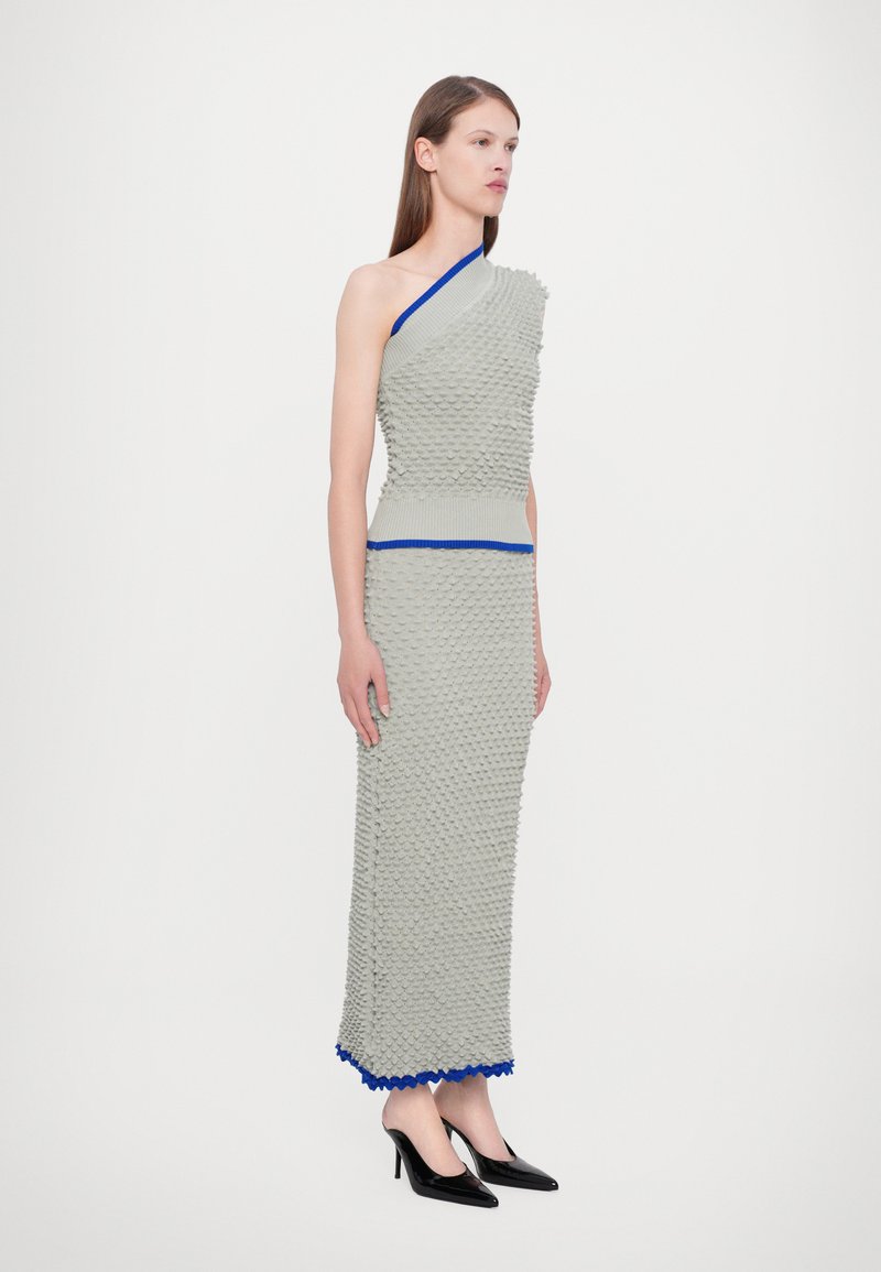 Grijze jurk met textuur en een asymmetrische halslijn, blauwe accenten op de taille en de zoom, gecombineerd met zwarte puntige hakken.