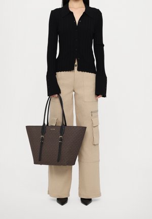 Persoon draagt een zwart langarmig, geknoopt shirt en beige cargobroek, houdt een grote bruine draagtas met zwarte handvatten vast.