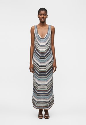 Γυναίκα που φοράει αμάνικο, πλεκτό φόρεμα μέχρι το πάτωμα με πολύχρωμες οριζόντιες ρίγες σε σχήμα ζιγκ-ζαγκ και μαύρα σανδάλια με ανοιχτά δάχτυλα, στέκεται μπροστά σε λευκό φόντο.